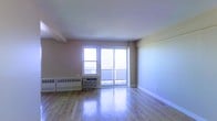 This single-floor space features a clean, open layout with light-toned hardwood flooring that reflects natural light throughout the room. The main living area is anchored by a large sliding glass door that leads to an outdoor balcony, accompanied by a fixed picture window beside it, together offering generous views of the surrounding urban skyline. The windows serve as the focal point of the space, drawing in abundant daylight and creating an airy, open atmosphere. Neutral-toned walls in warm beige complement the light wood flooring, contributing to a bright and welcoming environment. Along the base of the exterior wall, a wall-mounted heating unit provides climate control, supplemented by a window air conditioning unit, ensuring year-round comfort. White baseboards provide clean definition between the walls and flooring. The ceiling is smooth and well-lit by natural light, adding to the sense of openness. The overall layout is versatile and uncluttered, offering flexibility for a variety of furniture arrangements, making it a functional and well-lit living space with convenient outdoor access.