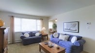 This single-floor residence offers a warm and inviting atmosphere designed for comfortable everyday living. The living area serves as the heart of the home, featuring large windows dressed with elegant curtains that allow generous amounts of natural light to fill the room, creating an airy and uplifting ambiance. The neutral-toned walls and light carpet complement the space, providing a versatile backdrop for a range of furnishings and décor styles. Tasteful wall art adds a finishing touch to the living area, enhancing its welcoming character. The living room connects openly to a dedicated dining area, which is visible through the space and benefits from its own window, bringing in additional natural light. The dining area is well-proportioned and provides a pleasant setting for meals. A track light fixture above ensures the dining space is well-lit. Throughout, the layout is thoughtfully designed to maximize flow and functionality, with clearly defined zones for relaxing and dining. The overall ambiance is calm, bright, and residential, making this a comfortable and practical space suited for a variety of lifestyles.