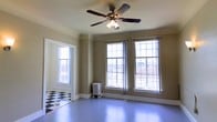 This single-floor space showcases a charming blend of classic architectural character and functional design. The main room is defined by an abundance of natural light streaming through multiple large, multi-pane windows that line the walls, offering views of the surrounding urban streetscape. The generous window arrangement creates an open, airy atmosphere throughout the day.

The flooring presents a notable contrast: a bold black-and-white checkered tile pattern anchors one section of the space, transitioning into smooth, painted hardwood floors in a soft blue-gray tone that extends across the main area. This combination adds visual interest and a touch of vintage charm.

A ceiling fan with a built-in light fixture is centrally mounted, providing both air circulation and supplemental lighting. The walls are finished in a warm, neutral earth tone, complemented by crisp white crown molding and baseboards that frame the room with traditional detailing.

A cast iron radiator positioned beneath the windows serves as both a functional heating element and a period-appropriate architectural accent. The overall ambiance of the space is one of understated elegance, with classic details and plentiful natural light making it a versatile and inviting environment.