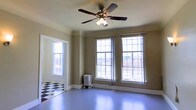 This single-floor space showcases a charming blend of classic architectural character and functional design. The main room is defined by an abundance of natural light streaming through multiple large, multi-pane windows that line the walls, offering views of the surrounding urban streetscape. The generous window arrangement creates an open, airy atmosphere throughout the day.

The flooring presents a notable contrast: a bold black-and-white checkered tile pattern anchors one section of the space, transitioning into smooth, painted hardwood floors in a soft blue-gray tone that extends across the main area. This combination adds visual interest and a touch of vintage charm.

A ceiling fan with a built-in light fixture is centrally mounted, providing both air circulation and supplemental lighting. The walls are finished in a warm, neutral earth tone, complemented by crisp white crown molding and baseboards that frame the room with traditional detailing.

A cast iron radiator positioned beneath the windows serves as both a functional heating element and a period-appropriate architectural accent. The overall ambiance of the space is one of understated elegance, with classic details and plentiful natural light making it a versatile and inviting environment.