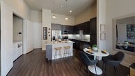This single-floor, one-bedroom, one-bathroom residence offers a thoughtfully designed open-concept layout that maximizes both functionality and style. The kitchen serves as a standout centerpiece, featuring dark espresso cabinetry that extends to impressive heights, granite countertops, a white subway tile backsplash, stainless steel appliances, and a central island with bar seating — ideal for casual dining or entertaining. Rich, dark hardwood flooring runs throughout the main living areas, complementing the soaring ceilings that lend a sense of grandeur to the space.

The living area is bathed in natural light thanks to expansive floor-to-ceiling windows and a glass-paneled door that opens directly onto a private balcony, offering outdoor views and seamless indoor-outdoor living. The bedroom continues the theme of generous natural light with large windows overlooking the balcony, providing a bright and airy retreat. Plush carpeting adds warmth underfoot.

The bathroom is finished with granite countertops, dark cabinetry, and neutral tile flooring, with direct access to a walk-in closet equipped with built-in shelving and hanging rods. A dedicated dining area adjacent to the kitchen completes this efficient and elegantly appointed home.