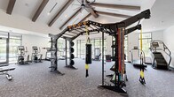 This well-appointed fitness center offers a generously sized, single-floor layout designed to accommodate a wide range of workout disciplines. The main workout area is defined by dramatic vaulted ceilings with exposed dark wood beams and recessed lighting, creating an open, energizing atmosphere. Large floor-to-ceiling windows and glass doors line the perimeter, flooding the space with natural light and providing views of surrounding greenery. The durable, textured carpet flooring throughout the primary area supports a comprehensive strength training zone equipped with power racks, cable machines, a pull-up and functional training rig, a heavy punching bag, free weights, and multiple benches. Cardio equipment includes treadmills, ellipticals, a stair climber, stationary bikes, and a rowing machine. An adjoining studio area features warm hardwood flooring, a ceiling fan, full-length wall mirrors, and distinctive birch-tree patterned wallcovering, making it well-suited for stretching, yoga, or low-impact training. This secondary space also provides direct outdoor access through sliding glass doors. Additional equipment storage, kettlebells, resistance bands, medicine balls, and a mounted display screen complete the facility's offerings, making it a comprehensive destination for fitness of all kinds.