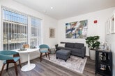Located at 2121 Wood St in Oakland, CA, this thoughtfully designed one-bedroom, one-bathroom unit offers a modern aesthetic with clean lines and contemporary finishes throughout. The single-floor layout is organized efficiently, beginning with an entry hallway adorned with framed artwork and louvered doors that set a polished tone upon arrival. Wide-plank wood-style flooring flows throughout, tying each room together seamlessly. The combined kitchen and living area forms the heart of the unit. The kitchen features sleek two-tone cabinetry with wood-grain upper cabinets and matte gray lower drawers, complemented by white countertops, a stainless steel sink, and stainless steel appliances including a range and built-in microwave. The living area is bright and inviting, with large windows that draw in natural light and provide street views. It is accented by bold abstract wall art and a neutral, comfortable seating arrangement. The bedroom is a cozy, well-appointed retreat featuring a frosted glass sliding door, a vibrant blue accent ceiling, recessed lighting, and framed photo wall art. The bathroom maintains the unit's modern character with a white countertop vanity, wood-toned cabinetry, light tile flooring, and clean white walls, creating a crisp, functional space.