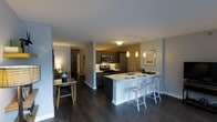 This single-floor residence offers a thoughtfully designed open-concept layout that seamlessly connects the kitchen, dining, and living areas. The kitchen is a standout feature, showcasing dark espresso cabinetry paired with light countertops, stainless steel appliances including a range, microwave, and refrigerator, and a large peninsula with a built-in sink. The peninsula doubles as a breakfast bar, accommodating modern metal bar stools that add an industrial-chic touch. Pendant lighting hangs above the peninsula, casting a warm ambiance that complements the recessed and track lighting overhead. Dark hardwood flooring runs throughout, grounding the space with a rich, contemporary feel. The open layout flows naturally into a dining nook, where a compact table and chairs are bathed in soft ambient light. A hallway visible from the main living area leads to additional rooms, hinting at a well-organized floor plan. The neutral gray walls and white trim provide a clean, modern backdrop that enhances the overall sense of openness. This space balances functional design with refined aesthetics, making it both practical and visually appealing.