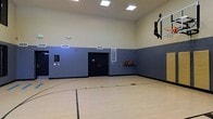 This indoor basketball facility is a purpose-built recreational space designed to support active play and athletic development. The court features a classic hardwood floor with clearly marked court lines, providing an authentic playing surface suited for both casual games and organized play. Soaring ceilings allow for full, unobstructed gameplay, and the space is illuminated by recessed overhead lighting that ensures bright, even coverage across the entire court. The walls are finished in a two-tone color scheme — a lighter upper half and a darker lower half — which is both aesthetically clean and practical for reducing glare. A wall-mounted basketball hoop is prominently positioned, accompanied by a bold silhouette graphic of a player dunking, adding a dynamic visual element to the space. A wall-mounted ball rack stocked with basketballs is conveniently accessible along the sideline, making equipment readily available for players. Entry to the court is provided through a set of sturdy double doors, and ventilation is maintained through wall-mounted HVAC vents. The overall design is functional and well-maintained, creating an inviting environment for players of all skill levels.
