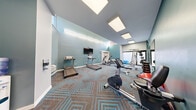 Located at 77 Village Way in Brookline, MA, this fitness center offers a thoughtfully designed exercise room that provides a comprehensive workout environment. The space is characterized by its striking teal-blue accent walls, crisp white ceilings, and a bold geometric-patterned carpet in brown and blue tones that adds a modern, energetic aesthetic throughout.

The room is generously equipped with cardio machines, including multiple treadmills and an elliptical trainer, as well as a stationary bike and a cable/weight machine with a dumbbell rack. Stability balls and a flat workout bench round out the strength and flexibility options. A wall-mounted flat-screen television is positioned for easy viewing during cardio sessions.

Natural light enters through clerestory-style windows positioned high along the walls, creating a bright and airy atmosphere without compromising privacy. Recessed panel lighting in the ceiling supplements the daylight to ensure the space is well-lit at all times. A mirrored wall section enhances the sense of openness and allows users to monitor their form.

Practical amenities include a cubby storage unit, a water dispenser, a wall-mounted HVAC unit, and hand sanitizer stations. The overall design balances functionality with a clean, modern ambiance, making it a well-appointed fitness facility for residents at 77 Village Way.
