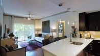 This single-floor residence showcases a thoughtfully designed open-concept layout that seamlessly connects the kitchen, dining, and living areas. The kitchen features a generous white countertop island with a modern undermount sink and sleek pendant lighting suspended from the ceiling, creating an elegant focal point. The transition into the living area is natural and effortless, where dark hardwood floors are complemented by a rich, patterned area rug. The living room is anchored by a tufted sofa and accent chairs arranged around a glass coffee table, with a large abstract artwork adorning the wall. A ceiling fan with integrated lighting adds both function and style overhead. Natural light filters in through a sliding glass door that leads to a private balcony overlooking lush greenery, enhancing the airy ambiance of the space. A dining area with upholstered chairs bridges the kitchen and living zones. Through a framed doorway, the bedroom is visible, featuring a neatly made bed and a decorative circular mirror, suggesting a well-finished and cohesive interior throughout the residence.