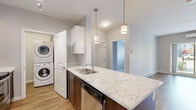This efficiently designed 1-bedroom, 1-bathroom residence delivers a clean, modern aesthetic across a single floor. The open-concept kitchen serves as the heart of the home, featuring a large island with striking marble-veined stone countertops, rich dark wood cabinetry, white upper cabinets with a classic subway tile backsplash, stainless steel appliances, and pendant lighting overhead. A convenient stacked washer and dryer unit is integrated directly into the kitchen area, maximizing functionality. The living room is bright and airy, finished with smooth hardwood-style flooring and a sliding glass door that opens to a private balcony with outdoor views, complemented by a wall-mounted air conditioning unit for year-round comfort. The bedroom offers a cozy carpeted floor, a built-in sliding-door closet with wire shelving, a baseboard heater, and a window overlooking the surrounding neighborhood. The bathroom continues the refined design language with marble-look stone countertops, a vessel sink, and clean tile flooring. A hallway connects the main living spaces with paneled white doors and warm wood-look flooring, tying the overall design together with a cohesive, contemporary feel.