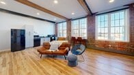 This 540 square foot apartment in Manchester, NH, seamlessly blends industrial charm with modern comfort. The open-concept living area showcases exposed brick walls and wooden ceiling beams, adding character and warmth to the space. Large windows flood the room with natural light, offering picturesque views of the surroundings.

The kitchen area, integrated into the main living space, features sleek black appliances and ample counter space. The living room is thoughtfully furnished with mid-century modern pieces, including a tan leather sofa and a blue accent chair, creating a cozy yet stylish atmosphere.

The bedroom, while compact, maximizes space with a queen-sized bed and built-in storage. A sliding barn-style door in blue adds a pop of color and separates the sleeping area from the main living space. The room is equipped with a wall-mounted air conditioning unit for comfort.

The bathroom is efficiently designed with a walk-in shower, modern fixtures, and a vanity with storage. Colorful accents in the shower curtain and bath mats add a playful touch to the otherwise neutral palette.

Throughout the apartment, warm wood flooring unifies the space and enhances the overall aesthetic. The combination of industrial elements, modern furnishings, and thoughtful design creates a unique and inviting urban living experience in this compact yet functional apartment.