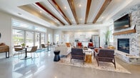 This thoughtfully designed single-floor amenity space offers a generous and versatile layout that seamlessly blends comfort, function, and contemporary style. The centerpiece of the space is a bright, open-concept great room featuring soaring ceilings adorned with rich, dark-stained wood beam accents and recessed lighting. Polished concrete floors reflect abundant natural light that pours in through expansive floor-to-ceiling windows and sliding glass doors, providing views of the surrounding outdoor areas. A striking floor-to-ceiling brick fireplace anchors the living area, adding warmth and architectural character.

The fully equipped kitchen is outfitted with crisp white shaker cabinetry, dark countertops, a subway tile backsplash, and stainless steel appliances, including a large French door refrigerator. Adjacent casual dining and seating areas create a welcoming, social atmosphere.

A dedicated fitness center offers a well-lit workout environment with large windows overlooking the outdoors, rubber flooring, cardio equipment including a treadmill, elliptical, and stationary bike, along with wall-mounted mirrors and a mounted television.

A sophisticated reception and lounge area features frosted glass partitions, curated artwork, and refined furnishings, providing an inviting and professional setting. Throughout, the space maintains a cohesive modern aesthetic with neutral tones and warm wood accents.