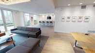 This single-floor amenity space is thoughtfully designed to balance comfort, functionality, and contemporary style. The open-plan layout flows seamlessly between distinct zones, unified by light wood-look flooring and a neutral white-and-gray color palette throughout.

The main lounge area is anchored by plush modular seating arrangements, including gray and navy sofas paired with leather accent chairs and low wooden coffee tables. A standout sculptural ceiling fixture—a geometric cluster of exposed bulbs set within a recessed tray—serves as a focal point, casting warm ambient light across the room. Floor-to-ceiling windows and glass doors invite abundant natural light and provide views of an outdoor terrace, creating an airy, open atmosphere.

A dedicated recreation zone features a ping-pong table and a vibrant abstract mural, adding a playful, energetic element to the space. Framed artwork displayed throughout the lounge contributes a gallery-like quality to the environment.

The kitchenette is well-appointed with dark stone countertops, white subway tile backsplash, a stainless steel sink with a commercial-style faucet, built-in microwave, under-counter refrigerator, and a wall-mounted television. A bar counter with tall stools and a mounted flat-screen TV provides additional casual dining and gathering space.