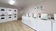 Located at 2625 Otranto Rd in North Charleston, South Carolina, this single-story community laundry facility is designed to provide a functional and accessible space for residents. The room features light wood-style vinyl flooring throughout, offering a clean and easy-to-maintain surface. Walls are finished in a neutral gray-white palette, contributing to a bright and orderly atmosphere.

The facility is equipped with a row of top-loading washing machines and stacked dryer units, offering ample capacity for multiple users simultaneously. Each machine features coin-operated or card-reader payment systems, and instructional signage is posted on the walls for user reference. A folding table is conveniently positioned within the space, providing a practical surface for sorting and folding laundry.

Overhead fluorescent lighting ensures the room is well-lit, while a window fitted with horizontal blinds allows for natural light and ventilation. Crown molding lines the upper perimeter of the walls, adding a subtle architectural detail. A dedicated entry door with a keypad or handle lock provides controlled access to the facility. A waste bin is also available for resident convenience. Overall, the space is straightforward, utilitarian, and well-organized to meet the everyday laundry needs of its users.