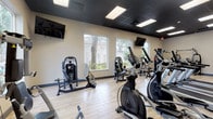 This fitness center is thoughtfully designed to accommodate a full range of workout styles within a single-level layout. The space is divided into distinct functional zones, each outfitted with professional-grade equipment. The cardio area features a lineup of treadmills, ellipticals, and stationary bikes from brands such as Spirit, arranged along windowed walls that bring in generous natural light and views of surrounding greenery. Adjacent to the cardio section, a strength training zone houses multiple cable machines, a lat pulldown station, and a functional trainer, providing comprehensive options for resistance-based workouts.

A dedicated free weights and functional fitness area offers a dumbbell rack, adjustable bench, stability balls, and resistance accessories, with motivational silhouette decals adorning the walls. This open zone benefits from additional natural light through large windows and provides ample floor space for stretching or bodyweight exercises. Wall-mounted flat-screen televisions are positioned throughout the facility for entertainment during workouts.

The flooring throughout consists of light wood-tone luxury vinyl planks, complemented by dark drop ceilings with recessed panel lighting that creates a focused, energetic atmosphere. Glass-panel entry doors and multiple exterior-facing windows ensure the space feels open and connected to the outdoors.