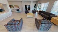 Located in Wilmer, Texas, this single-story community clubhouse at Liberty Crossing offers a well-appointed gathering space designed for comfort and functionality. The open-concept layout is anchored by large-format light tile flooring throughout, creating a clean and cohesive aesthetic across all areas.

The spacious main lounge area is furnished with colorful patterned accent chairs and a sleek dark leather sofa, arranged around a central coffee table. Natural light pours in through large windows fitted with white horizontal blinds, and a set of glass double doors provides direct access to an outdoor pool area. Vibrant abstract artwork adorns the neutral gray walls, adding a lively, welcoming character to the space.

Adjacent to the lounge is a casual dining and work area featuring light wood tables paired with modern teal and black chairs. A bold, colorful painting of a tree serves as a focal point on the accent wall, and built-in cabinetry below provides additional storage.

The kitchen is outfitted with dark espresso cabinetry, granite countertops, stainless steel appliances including a full-size refrigerator, microwave, and dishwasher, and a large island with bar seating. Recessed lighting throughout the space ensures bright, even illumination. Decorative touches, including Texas-themed wall art and potted plants, contribute warmth and personality to this thoughtfully designed community hub.