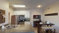 This single-floor residence offers a thoughtfully designed open-concept layout that seamlessly connects the kitchen, dining, and living areas. The kitchen is outfitted with stainless steel appliances — including a refrigerator, electric range, and over-the-range microwave — complemented by crisp white cabinetry, neutral-toned countertops, and a peninsula with an integrated sink that doubles as a casual breakfast bar. The dining area sits adjacent to the kitchen and provides ample space for entertaining. The living room is warm and inviting, featuring crown molding, wood-style flooring, and a sliding glass door that fills the space with natural light and leads to an outdoor patio with views of the surrounding area. The bedroom offers plush carpeting, a window with vertical blinds for light control, and a wall-mounted television setup. The full bathroom includes a tub and shower combination with tile surround and wood-style flooring. A dedicated laundry area with a washer is also visible within the unit. Throughout the residence, neutral wall tones and consistent finishes create a cohesive, modern aesthetic that is both functional and welcoming.