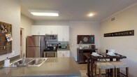 This single-floor residence offers a thoughtfully designed open-concept layout that seamlessly connects the kitchen, dining, and living areas. The kitchen is outfitted with stainless steel appliances — including a refrigerator, electric range, and over-the-range microwave — complemented by crisp white cabinetry, neutral-toned countertops, and a peninsula with an integrated sink that doubles as a casual breakfast bar. The dining area sits adjacent to the kitchen and provides ample space for entertaining. The living room is warm and inviting, featuring crown molding, wood-style flooring, and a sliding glass door that fills the space with natural light and leads to an outdoor patio with views of the surrounding area. The bedroom offers plush carpeting, a window with vertical blinds for light control, and a wall-mounted television setup. The full bathroom includes a tub and shower combination with tile surround and wood-style flooring. A dedicated laundry area with a washer is also visible within the unit. Throughout the residence, neutral wall tones and consistent finishes create a cohesive, modern aesthetic that is both functional and welcoming.