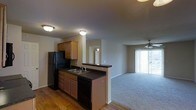 This single-story unit features an open-concept layout that seamlessly connects the kitchen and living area. The kitchen is equipped with medium-toned wooden cabinetry, dark countertops, a double-basin sink, and black appliances including a full-sized refrigerator and dishwasher. A peninsula counter serves as a natural divider between the kitchen and the adjoining living space, offering additional prep or seating space while maintaining an open feel. The kitchen area features durable hardwood-style flooring, while the living area transitions to plush carpeting, creating a clear distinction between the two zones. The living room is bathed in natural light streaming through sliding glass doors that open to what appears to be a balcony or patio, fitted with vertical blinds for privacy control. A ceiling fan is centrally positioned in the living area, providing both lighting and air circulation. A hallway visible from the kitchen leads to additional areas of the unit. Flush-mount ceiling lights illuminate both the kitchen and living areas, contributing to the overall bright and functional ambiance of the space.