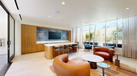 Located at 1650 Lincoln Blvd in Santa Monica, California, this thoughtfully designed single-floor lounge space offers a refined and welcoming atmosphere with a strong emphasis on modern aesthetics and functionality. The open-plan layout maximizes every square foot, creating a seamless flow between distinct functional zones.

At the heart of the space is a sleek kitchenette and bar area, anchored by a rounded island with a crisp white quartz countertop and warm natural wood cabinetry. A textured blue-toned accent wall frames a wall-mounted flat-screen display, adding visual depth and a contemporary focal point. Built-in cabinetry and integrated appliances keep the design clean and cohesive.

The living area is furnished with an eclectic yet harmonious mix of curved leather seating, sculptural accent chairs in rich cognac and teal blue upholstery, and a boldly patterned area rug in earthy tones. A tucked-away dining nook with banquette seating and pendant lighting provides an intimate setting within the larger space.

Floor-to-ceiling glass walls and doors flood the interior with natural light and offer direct views of lush outdoor greenery and a patio area. Light-toned flooring throughout enhances the bright, airy ambiance. This space is ideal as a communal lounge or amenity room, combining comfort, style, and a distinctly California sensibility.