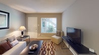 This single-floor unit offers a practical and comfortable layout designed to maximize its efficient footprint. The living room serves as the central gathering space, featuring wall-to-wall carpeting, neutral gray walls, and a large window with blinds that allows natural light to filter in while offering a view of the surrounding greenery. The open entry door adds accessibility and a welcoming feel to the area.

The kitchen is thoughtfully designed with white cabinetry extending to the upper walls, providing generous storage space. Laminate countertops run the length of the galley-style kitchen, which includes a stainless steel sink, a dishwasher, and a full-sized refrigerator. Wood-look flooring adds a warm contrast to the white cabinetry.

The bedroom is a well-sized, carpeted retreat featuring multiple windows that bring in natural light and outdoor views. Two closets offer substantial storage, including one with a rod and shelf system. The textured ceiling is consistent throughout the unit.

The bathroom features tile wainscoting, a shower and tub combination with a decorative curtain, and a vanity visible from the bedroom hallway, completing this straightforward and functional layout.