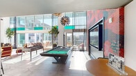 Located at 132 Unity St in Irvine, CA, this striking single-floor communal amenity space is designed to impress with bold aesthetics and a lively, social atmosphere. Soaring double-height ceilings and expansive floor-to-ceiling glass walls flood the interior with abundant natural light while providing sweeping views of the surrounding outdoor terrace and beyond.

The game room area anchors the space with a sleek pool table and a foosball table positioned on polished large-format tile flooring, complemented by a dramatic sputnik-style chandelier overhead. A large wall-mounted display screen is set against a vivid, oversized floral mural in rich reds and pinks, serving as a bold artistic focal point.

Adjacent to the game area, a stylish lounge features eclectic floral-upholstered armchairs arranged around a round coffee table, accented by a graphic black-and-white lattice wall installation above. A fully equipped wet bar with bar seating provides a social gathering spot, finished with dark cabinetry and tile detailing.

Lush indoor potted trees add organic warmth throughout, while sheer curtains soften the glass perimeter. Sliding glass doors open directly onto a furnished outdoor terrace, seamlessly blending indoor and outdoor living. The overall design combines contemporary architecture with vibrant, curated décor for an energetic and welcoming communal environment.