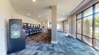 This single-floor entertainment lounge is designed to offer a well-rounded recreational experience across a generously proportioned open layout. The space is divided into distinct activity zones that flow seamlessly into one another, all unified by patterned multi-tone carpet in shades of blue, brown, and gray that adds visual depth underfoot.

One wing of the space is dedicated to an impressive lineup of pinball machines arranged along the wall, complemented by high-top bar seating nearby for spectators. Recessed lighting illuminates this area, and the neutral-toned walls and clean white ceilings contribute to a modern, polished aesthetic.

The adjacent gaming area serves as the centerpiece of the lounge, featuring a full-size pool table and a foosball table. Floor-to-ceiling windows along multiple walls flood this zone with natural light and provide views of the surrounding outdoor greenery. Textured geometric wall panels in soft sage tones add a sophisticated design element, while a wall-mounted television and plush teal lounge chairs create a comfortable viewing nook.

A wet bar with cabinetry provides a convenient amenity, and additional high-top tables with swivel stools offer casual seating throughout. The overall ambiance balances energetic recreation with contemporary design, making it a versatile and inviting communal destination.