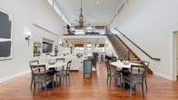 Located at 29 French Mill Run in Cuyahoga Falls, Ohio, this beautifully appointed community clubhouse is a welcoming gathering space defined by its impressive architectural character. Upon entry, soaring double-height ceilings draw the eye upward to a dramatic chandelier and an upper-level mezzanine with wrought iron railings, accessible via a graceful staircase. Rich hardwood flooring flows throughout, anchoring the bright, open layout. The main gathering area offers generous seating arrangements with dining tables and lounge furniture, bathed in natural light from large windows dressed with bamboo Roman shades. A dedicated conference or dining room provides a more intimate setting, featuring an oversized table, a wall-mounted TV, and broad windows overlooking the surrounding community. The kitchenette is outfitted with white cabinetry, granite countertops, a sink, and a coffee station, offering residents convenient refreshment amenities. A recreation area features a pool table alongside a built-in entertainment center and comfortable seating. The space also includes accessible restrooms finished with granite vanities, tile flooring, and ADA-compliant fixtures. Thoughtfully designed and meticulously maintained, French Mill Run's clubhouse delivers a refined yet comfortable environment for residents to socialize and relax.