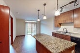 This thoughtfully designed one-bedroom, one-bathroom residence offers an efficient and comfortable layout across a single floor. The open-concept kitchen serves as the heart of the home, showcasing rich warm-toned cabinetry, a granite countertop island with pendant lighting overhead, and a full suite of black appliances including a refrigerator, range, and microwave. Track lighting complements the pendants, illuminating the space effectively. The kitchen flows seamlessly into the living area, which is finished with hardwood-style flooring and leads to a sliding glass door that opens onto a private balcony, allowing natural light to fill the space. The bedroom features plush carpeting, a ceiling fan, and a window with views of the surrounding neighborhood. Warm cherry-toned doors throughout lend a cohesive, polished aesthetic. The bathroom is equipped with a granite-topped vanity, a tub-shower combination, and a toilet, with convenient access to an in-unit stacked washer and dryer. A hallway and dedicated closet provide practical storage solutions. Rich wood trim accents and consistent finishes throughout create a unified and inviting atmosphere.