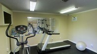 This fitness center offers a practical and functional space designed to support a well-rounded workout routine. The room is finished with neutral, light-colored walls and a textured ceiling, giving it a clean and utilitarian feel. Overhead fluorescent lighting ensures the space is well-lit throughout, creating an energizing atmosphere for exercise.

The floor is covered in dark rubber matting, providing both traction and durability suited for high-impact activities. A standout feature of the room is the large wall-mounted mirror panel, which spans a significant portion of one wall, visually expanding the space and allowing users to monitor their form during workouts.

Cardio equipment is prominently featured, including a treadmill and an elliptical machine, both positioned for ease of use with ample surrounding clearance. Additional strength and conditioning equipment is visible in the mirror's reflection, suggesting a broader range of training options within the space. A security camera is also present, contributing to a safe and monitored environment.

Overall, this fitness center combines essential amenities with a straightforward, no-frills design, making it a convenient and accessible facility for daily exercise.