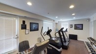 This fitness room offers a focused cardio workout environment featuring a selection of Life Fitness brand equipment, including elliptical trainers and treadmills. The space is finished with neutral white walls and recessed lighting throughout the ceiling, ensuring consistent, bright illumination across the room. Windows fitted with white horizontal blinds allow for natural light while maintaining privacy. The flooring features a low-pile carpet suited for exercise use. Wall-mounted informational signage and framed artwork provide subtle visual interest. A water cooler station is conveniently positioned near the equipment. The room maintains a clean, functional layout, with equipment arranged to allow comfortable access and circulation. The overall ambiance is practical and tidy, designed to support a straightforward cardio fitness routine in a well-maintained setting.