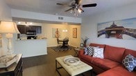 This single-floor residence offers an open-concept layout that seamlessly connects the living, dining, and kitchen areas, creating a cohesive and functional living environment. The living room serves as the central gathering space, featuring a ceiling fan with integrated lighting, a textured ceiling, and rich dark-toned flooring that grounds the room with warmth. A bold rust-colored sofa with decorative throw pillows anchors the seating area, complemented by a tufted ottoman that doubles as a coffee table. Wall art adds personality and a creative flair to the space. The dining area sits just beyond the living room, defined by a classic chandelier overhead and a round dining table with Windsor-style chairs set atop a graphic black-and-white area rug. The kitchen is accessible via a raised breakfast bar counter, offering a convenient pass-through that keeps the space open and social. Dark cabinetry and black appliances provide a sleek contrast to the light countertops and neutral walls. Throughout the home, the consistent flooring, warm neutral palette, and thoughtful decor create a cohesive, inviting ambiance suited for both everyday living and entertaining.
