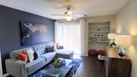 This well-appointed single-floor space offers a thoughtfully designed interior that balances comfort and style. The living room serves as the centerpiece of the home, featuring rich hardwood-style flooring that anchors the space with warmth and sophistication. A bold accent wall in deep slate blue adds a striking visual contrast against the neutral tones of the room, complemented by a large piece of wall art that enhances the curated aesthetic.

The living area is furnished with a plush, tufted sectional sofa adorned with decorative pillows, creating an inviting and relaxed atmosphere. A glass-top coffee table sits atop a patterned area rug, adding elegance to the space. Built-in white shelving along one wall provides both functional storage and a platform for tasteful décor displays.

Natural light floods the room through a large sliding glass door dressed with sheer white curtains and vertical blinds, offering a glimpse of the outdoor surroundings. A ceiling fan with an integrated light fixture ensures year-round comfort. Accent ottomans in a warm terracotta hue add a pop of color and additional seating, tying the design together into a cohesive and welcoming living environment.