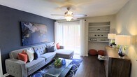 This well-appointed single-floor space offers a thoughtfully designed interior that balances comfort and style. The living room serves as the centerpiece of the home, featuring rich hardwood-style flooring that anchors the space with warmth and sophistication. A bold accent wall in deep slate blue adds a striking visual contrast against the neutral tones of the room, complemented by a large piece of wall art that enhances the curated aesthetic.

The living area is furnished with a plush, tufted sectional sofa adorned with decorative pillows, creating an inviting and relaxed atmosphere. A glass-top coffee table sits atop a patterned area rug, adding elegance to the space. Built-in white shelving along one wall provides both functional storage and a platform for tasteful décor displays.

Natural light floods the room through a large sliding glass door dressed with sheer white curtains and vertical blinds, offering a glimpse of the outdoor surroundings. A ceiling fan with an integrated light fixture ensures year-round comfort. Accent ottomans in a warm terracotta hue add a pop of color and additional seating, tying the design together into a cohesive and welcoming living environment.