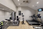 Located at 1001 Pinemont Drive in Houston, TX, this dedicated fitness center offers a thoughtfully designed workout environment across a single floor. The space features vaulted ceilings that lend an open, airy feel, complemented by recessed lighting and neutral gray walls. Light wood-style flooring covers the main areas, while rubber mats are positioned beneath equipment for safety and durability.

The gym is divided into functional zones. The cardio area is equipped with treadmills, elliptical trainers, and a stationary bike, all positioned near a windowed entry door that allows natural light to filter in. A wall-mounted flat-screen TV adds convenience for cardio sessions. The strength training zone features a multi-functional cable machine, a leg press/extension station, a flat bench, and a rack of dumbbells for free weight exercises. A floor mat guide is mounted to the wall to assist with stretching routines.

A mirrored section expands the sense of space and provides a dedicated area for functional training, complete with medicine balls and additional cable attachments. Built-in white cabinetry near the ceiling offers storage, while a posted fitness center rules sign reflects a structured, community-oriented environment. A ceiling fan and ventilation system ensure comfort during workouts, making this gym a comprehensive and practical fitness facility.