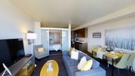 This open one-bedroom, one-bathroom unit is thoughtfully designed to maximize every square foot of its compact footprint. The living area serves as the heart of the space, featuring large windows that invite abundant natural light and provide views of the surrounding area. A sliding glass partition wall separates the sleeping quarters from the main living space, offering privacy while maintaining an airy, open feel throughout. The bedroom accommodates a full bed and benefits from the natural light that filters through the glass panels.

The kitchen is efficiently laid out along one wall, featuring dark wood cabinetry, stainless steel appliances including a microwave, range, and dishwasher, and a light-toned countertop with a single-basin sink. A compact dining area positioned nearby provides space for casual meals.

The living room offers enough space for a full sofa, armchair, and media console, creating a comfortable lounge area. Elevated ceilings contribute to a sense of spaciousness throughout. A private balcony accessible from the living area extends the usable space outdoors, ideal for taking in surrounding views.