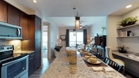 This single-floor residence showcases a thoughtfully designed open-concept layout that seamlessly connects the kitchen, dining, and living areas. The kitchen is anchored by a generous granite-topped island with striking natural veining, complemented by dark cabinetry that provides a bold contrast against the light walls and wood-tone flooring. Pendant lights with decorative cylindrical shades hang above the island, adding an elegant focal point to the space.

The dining area transitions naturally into the living room, where a ceiling fan with an integrated light fixture maintains airflow and comfort. Large floor-to-ceiling windows dressed with layered draping allow abundant natural light to pour in, while offering views of an outdoor balcony beyond. The living area is spacious and versatile, accommodating a variety of seating arrangements. A hallway visible from the kitchen leads to additional rooms, suggesting a well-organized and functional floor plan. The overall ambiance is modern yet warm, combining rich materials with neutral tones for a refined, comfortable atmosphere.