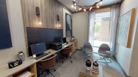 This single-floor business center in Fort Worth, TX (76109) is a thoughtfully designed shared workspace that balances functionality with refined contemporary aesthetics. The space is organized into distinct zones, each serving a specific professional purpose while maintaining a cohesive design language throughout.

The main workstation area features a built-in counter-height desk spanning the length of one wall, finished in light wood cabinetry with open shelving below. Two computer workstations with monitors, a shared printer, and ample desk space accommodate multiple users simultaneously. Warm pendant globe lights and wall sconces mounted against a richly textured wood-paneled accent wall provide focused task lighting, while bold circular-patterned artwork in navy and white adds visual interest.

A private office nook, accessible through a glass-paneled gray door, offers a quieter individual workspace with its own dedicated monitor, leather task chair, and the same warm wood cabinetry. A glass-enclosed conference or meeting room, visible from the corridor and accessible via French-style doors, provides a more private setting for collaborative work.

The lounge area features two upholstered swivel chairs, a low wood coffee table with decorative accessories, and a large floor-to-ceiling window dressed with sheer curtains that invites natural light and outdoor views. An eye-catching sculptural branch chandelier overhead ties the entire space together with an elevated, modern flair.