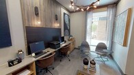 This single-floor business center in Fort Worth, TX (76109) is a thoughtfully designed shared workspace that balances functionality with refined contemporary aesthetics. The space is organized into distinct zones, each serving a specific professional purpose while maintaining a cohesive design language throughout.

The main workstation area features a built-in counter-height desk spanning the length of one wall, finished in light wood cabinetry with open shelving below. Two computer workstations with monitors, a shared printer, and ample desk space accommodate multiple users simultaneously. Warm pendant globe lights and wall sconces mounted against a richly textured wood-paneled accent wall provide focused task lighting, while bold circular-patterned artwork in navy and white adds visual interest.

A private office nook, accessible through a glass-paneled gray door, offers a quieter individual workspace with its own dedicated monitor, leather task chair, and the same warm wood cabinetry. A glass-enclosed conference or meeting room, visible from the corridor and accessible via French-style doors, provides a more private setting for collaborative work.

The lounge area features two upholstered swivel chairs, a low wood coffee table with decorative accessories, and a large floor-to-ceiling window dressed with sheer curtains that invites natural light and outdoor views. An eye-catching sculptural branch chandelier overhead ties the entire space together with an elevated, modern flair.