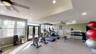 This fitness center is thoughtfully designed to support a comprehensive workout experience within a single, open floor layout. The space is finished with neutral-toned carpet flooring and sage green walls, creating a calm, focused atmosphere conducive to exercise.

The cardio zone is anchored near large windows and French doors that invite abundant natural light and outdoor views. It features multiple treadmills, elliptical trainers, and stationary cycling bikes, all positioned to take advantage of the bright, airy environment. Ceiling fans overhead help maintain comfortable airflow throughout the space.

The strength training area is well-appointed with a multi-station cable machine, a dedicated weight bench, and a full rack of dumbbells spanning a wide range of weights. Medicine balls and a foam roller are also available, supporting functional and flexibility training. A wall-mounted exercise mat adds further versatility for floor-based workouts.

Full-length mirrors line a portion of the walls, enhancing the sense of space while allowing users to monitor their form during exercises. Recessed lighting supplements the natural light, ensuring the room remains well-lit at all times. Informational boards are posted on the walls, providing guidance and fitness center policies. The overall design is clean, practical, and welcoming, making this fitness center a well-rounded amenity for daily use.