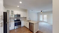 This single-story residence is designed with an open-concept layout that seamlessly connects the kitchen and living area, creating a cohesive and functional living space. The kitchen is a standout feature, appointed with crisp white shaker-style cabinetry, light granite countertops, a subway tile backsplash, and a full suite of stainless steel appliances including a range, microwave, refrigerator, and dishwasher. A peninsula with an undermount sink and additional counter space extends into the main living area, providing both a practical workspace and a natural dividing point between the kitchen and living room. Hardwood-style flooring runs throughout the kitchen and entry areas, while plush carpeting carries through the living room and bedroom. The living area benefits from natural light through a set of glass-paneled doors and is fitted with a ceiling fan overhead. Crown molding and recessed lighting add refined detail to the shared spaces. The bedroom is a calm, neutral retreat featuring carpeted flooring and a reach-in closet with built-in shelving for organized storage. The bathroom is finished with a granite-topped vanity, hardwood-style flooring, a tub-shower combination with tiled surround, and a window that introduces natural light.