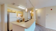 This single-story residence is designed with a practical, open-concept layout that connects the kitchen and living areas seamlessly. The kitchen is equipped with light maple cabinetry, a white electric range with overhead ventilation, a full-size refrigerator, and generous countertop workspace. A peninsula with an integrated double sink extends into the living area, offering additional prep space and casual seating potential. Pendant lighting and track lighting illuminate the kitchen, while a single hanging fixture anchors the adjacent living area, which features neutral carpet flooring throughout.

The bedrooms are carpeted and finished in warm neutral tones, each with windows fitted with horizontal blinds that bring in natural light and provide views of the surrounding greenery and a water feature. One bedroom includes direct access to a private bathroom and a separate closet. The bathrooms feature light maple vanities with countertop sinks, large wall-mounted mirrors with strip lighting, and tile flooring. A separate tub and shower area with white tile surrounds is also present. The overall color palette is consistently light and neutral, contributing to a clean, functional ambiance throughout the space.