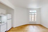 Located at 1030 Post St in San Francisco's Lower Nob Hill neighborhood, this one-bedroom, one-bathroom unit offers a clean, minimalist aesthetic throughout its efficiently laid-out single floor. The living area greets you with warm hardwood flooring, crisp white walls, and a multi-pane window that frames views of the surrounding urban streetscape while drawing in natural light. A baseboard heating unit beneath the window ensures year-round comfort. A compact kitchen is integrated into the main living space, featuring white cabinetry, a sink, and essential appliances including a refrigerator. A narrow hallway with continuous hardwood flooring connects the main living area to the private quarters, maintaining a cohesive design flow throughout. The bedroom is carpeted for added comfort and includes its own window with a street-facing view and baseboard heating, providing a quiet and cozy retreat. The full bathroom is finished in all-white, featuring a vanity with a mirrored medicine cabinet, toilet, and a bathtub with a shower — all accented by chrome fixtures and bright overhead lighting. The unit's consistent use of white walls, light wood flooring, and natural light creates a bright, clean atmosphere suited for urban living in the heart of San Francisco.
