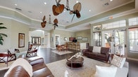 This expansive single-story space exhibits a refined, tropical-influenced design characterized by soaring tray ceilings adorned with dramatic multi-blade ceiling fans featuring woven rattan blades. The interior is unified by warm sage-green walls, crisp crown molding, and rich dark hardwood flooring throughout, creating a sophisticated yet welcoming atmosphere.

The main gathering area is generously proportioned, accommodating multiple seating arrangements including leather sofas and armchairs anchored by large area rugs. Recessed lighting complements an abundance of natural light streaming through numerous windows, including arched transom windows that frame outdoor greenery. French doors provide additional connectivity to exterior areas.

A bar-height counter with granite countertops serves as both a functional and social focal point, equipped with bar seating, dark cabinetry, a refrigerator, and small appliances. Framed artwork accents the surrounding walls. An adjacent dining area with wooden chairs and tables offers additional seating.

A quieter lounge area near the windows features comfortable seating and a rounded ottoman, ideal for relaxed conversation. Separate rooms accessible through double French doors offer additional flexible space, furnished with accent chairs and side tables for more intimate gatherings.