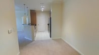 This single-floor space features an open-concept design that integrates the living area and kitchen into one cohesive layout. The living area is finished with neutral-toned carpet, lending warmth and comfort underfoot, while the kitchen and adjoining hallway transition to light-colored ceramic tile flooring. The kitchen is equipped with black appliances, including a full-sized refrigerator and range, and features a raised breakfast bar with a dark countertop that serves as both a functional workspace and a casual dining area. Pendant lighting hangs above the bar, adding focused illumination and a touch of style. A track lighting fixture in the hallway provides additional ambient light throughout the central corridor. The walls are painted in a soft, neutral beige tone throughout, contributing to a clean and airy atmosphere. A paneled interior door at the end of the hallway leads to additional areas of the unit. Crown molding accents are visible, adding a subtle architectural detail. The overall layout is practical and well-organized, making efficient use of the available space.