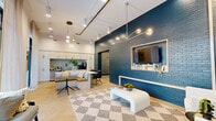 This single-floor amenity space is thoughtfully designed to serve as a communal hub, combining a fully equipped kitchen with a stylish lounge and game room. The layout flows seamlessly between zones, unified by light herringbone-patterned flooring and soaring ceilings that lend an airy, open quality throughout.

The kitchen is outfitted with floor-to-ceiling white cabinetry, a light blue subway tile backsplash, stainless steel appliances, a water station, and a marble-topped island with seating. Sheer drapery filters natural light from tall windows, enhancing the bright, clean aesthetic.

The lounge area serves as the social centerpiece, anchored by a dramatic deep teal brick accent wall housing a built-in media display. A sculptural multi-globe chandelier and track lighting illuminate the space, while a checkered area rug and modular seating with gold accent pillows add warmth and personality.

Adjacent to the lounge, the game room features a full-size pool table beneath a coffered ceiling with recessed pendant lights. Teal-painted double doors and a textured brick partition wall continue the cohesive color story throughout. Together, these spaces create a vibrant, design-forward environment ideal for gathering and recreation.