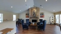 This single-floor space exudes a warm and inviting atmosphere, thoughtfully designed for comfortable gathering and relaxation. The focal point of the main lounge area is a floor-to-ceiling brick fireplace, framed by a large figurative artwork and flanked by bold Americana-themed wall pieces that lend the space a distinctive, patriotic character. Vaulted ceilings rise dramatically overhead, enhancing the open, airy feel of the room while recessed lighting provides a clean, well-lit environment throughout. Rich, dark hardwood flooring runs the length of the space, adding warmth and visual continuity. A seating arrangement of navy blue wingback and accent chairs surrounds a wooden coffee table, creating an intimate conversation area in front of the fireplace. Adjacent to the main lounge, a casual dining or bar area features high-top tables and stools positioned near a window that allows natural light to filter in. The overall design balances rustic and contemporary elements, combining exposed brick, warm wood tones, and curated artwork to create a cohesive and welcoming environment suited for both social gatherings and quiet relaxation.