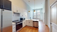 This thoughtfully designed single-floor unit offers a clean, modern aesthetic throughout. The kitchen serves as the heart of the space, showcasing tall ceilings that amplify the sense of openness. White shaker-style cabinetry with matte black hardware lines the walls, complemented by a classic white subway tile backsplash. A full suite of stainless steel appliances includes a side-by-side refrigerator with an in-door dispenser, an electric range, a built-in microwave, and a dishwasher. Two elegant brass pendant lights hang above the kitchen peninsula, which features a single-basin sink with a matte black faucet and light-toned countertops. Warm hardwood-style flooring flows seamlessly throughout the living areas and kitchen, adding richness and continuity to the space. Large windows dressed with horizontal blinds invite generous natural light into both the kitchen and adjoining rooms, offering views of the surrounding exterior. A dedicated laundry room provides added convenience, housing a full-size front-loading washer and dryer alongside built-in wire shelving for storage. The bathroom is finished with large-format dark tile flooring, a white vanity with a framed mirror, and accessibility-friendly grab bars, reflecting a practical and inclusive design approach.