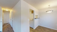 This single-story unit offers a straightforward and functional layout with two bedrooms and two full bathrooms. The living area greets residents at the front entry, where large windows allow generous natural light to fill the space, complemented by wood-style plank flooring and a textured ceiling. A pendant light fixture adds a subtle decorative touch near the entryway.

The kitchen is accessible via a pass-through opening from the main living area, featuring dark laminate countertops, white cabinetry with warm wood trim accents, a dishwasher, and wood-style flooring that carries through from the main living space. A breakfast bar-style counter along the pass-through provides convenient additional workspace.

Both bedrooms are carpeted and well-proportioned, each receiving natural light through windows fitted with horizontal blinds. The neutral wall tones throughout contribute to a clean, move-in-ready atmosphere.

The two bathrooms include wood-style flooring, tub-shower combinations with brushed nickel hardware, and vanities with dark countertops and lighted mirrors. The overall design is practical and cohesive, with consistent finishes maintained throughout the unit.