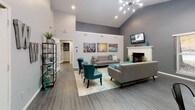 This single-floor community clubhouse is designed to serve as a welcoming social hub, combining modern aesthetics with functional spaces. The main lounge area commands attention with its soaring vaulted ceilings and a dramatic sputnik chandelier overhead. Neutral gray walls are complemented by bold teal accent chairs and a contemporary gray sofa arranged around a classic white fireplace. Vibrant cityscape artwork adds visual interest, while dark hardwood-style flooring runs throughout, grounding the space with a sleek, cohesive look.

Adjacent to the lounge, a dedicated dining area provides flexible seating with multiple round tables and industrial-style metal chairs. Large windows fitted with horizontal blinds line the walls, flooding the room with natural light and offering views of surrounding greenery. A glass-paned exterior door provides direct outdoor access, enhancing the open, airy atmosphere.

The fully equipped kitchen features dark espresso cabinetry paired with stainless steel appliances, including a range, microwave, and French door refrigerator. Light-toned flooring and a breakfast bar with bar stool seating make it practical for community gatherings and events. A dropped ceiling with panel lighting ensures the kitchen remains bright and functional. Together, these spaces create an inviting, well-appointed environment suited for both casual socializing and organized community activities.