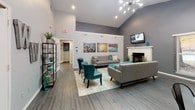 This single-floor community clubhouse is designed to serve as a welcoming social hub, combining modern aesthetics with functional spaces. The main lounge area commands attention with its soaring vaulted ceilings and a dramatic sputnik chandelier overhead. Neutral gray walls are complemented by bold teal accent chairs and a contemporary gray sofa arranged around a classic white fireplace. Vibrant cityscape artwork adds visual interest, while dark hardwood-style flooring runs throughout, grounding the space with a sleek, cohesive look.

Adjacent to the lounge, a dedicated dining area provides flexible seating with multiple round tables and industrial-style metal chairs. Large windows fitted with horizontal blinds line the walls, flooding the room with natural light and offering views of surrounding greenery. A glass-paned exterior door provides direct outdoor access, enhancing the open, airy atmosphere.

The fully equipped kitchen features dark espresso cabinetry paired with stainless steel appliances, including a range, microwave, and French door refrigerator. Light-toned flooring and a breakfast bar with bar stool seating make it practical for community gatherings and events. A dropped ceiling with panel lighting ensures the kitchen remains bright and functional. Together, these spaces create an inviting, well-appointed environment suited for both casual socializing and organized community activities.