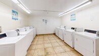 Located at 1250 N June St in Los Angeles, this shared laundry facility is a single-room space designed to serve the practical needs of building residents. The room is laid out efficiently, housing a generous bank of commercial-grade top-loading washing machines along one wall and a row of standalone dryers along the opposite side, providing ample capacity for multiple users at once. The space is finished with durable, easy-to-clean tan tile flooring throughout, and the white-painted walls and cinder block accents give the room a clean, utilitarian appearance. Overhead fluorescent lighting ensures the space is well-lit at all times. Two folding tables are positioned within the room, offering convenient surfaces for sorting and folding laundry. Informational signage is posted on the walls, including usage guidelines and etiquette reminders for shared use. The layout allows for comfortable movement between machines and folding areas. Overall, this laundry facility is a functional, well-equipped communal space designed with practicality and ease of use in mind, serving as a valuable shared amenity for residents of the property.